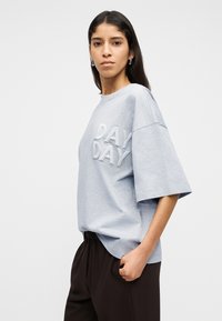 Γυναίκα με μακριά σκούρα μαλλιά στέκεται με το χέρι στην τσέπη, φορόντας ανοιχτό γκρι φαρδιά μπλούζα με την επιγραφή «DAY DAY» και σκούρο παντελόνι, με φόντο λευκό.