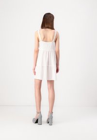 Robe blanche sans manches avec un corsage froncé et une jupe à volants. Les caractéristiques notables incluent des bretelles ajustables et un tissu doux et texturé.