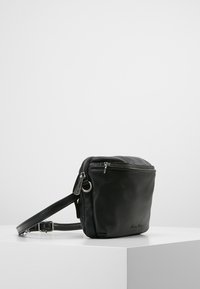 Mustassa nahkaisessa olkalaukussa on teksturoidun pinnan, vetskarisulku, säädettävä hihna ja hopeanväriset yksityiskohdat. Kompaktin ja neliömäisen muotoilun.