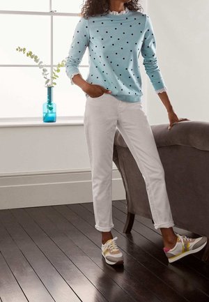 Femme en pull bleu clair à pois, pantalon blanc et baskets colorées, debout à côté d'un canapé gris sur un sol en bois foncé.