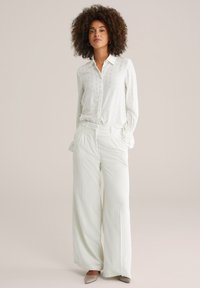 Blusa bianca leggera e con bottoni, decorata con motivi discreti, abbinata a pantaloni a gamba larga e plissettati di colore crema. Tessuto morbido e superficie liscia.