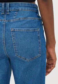 Plave traperice s visokim strukom, stražnjim džepovima i glatkom teksturom s kontrastnim šavovima.