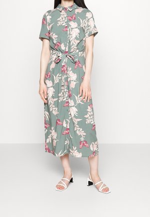 Robe chemise midi verte avec imprimés floraux roses et papillons, manches courtes, ceinture à la taille, portée avec des sandales blanches à talons.