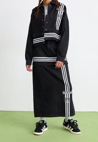 Svart denimjacka i croppad modell med vita ränder, lagd över en lång svart kjol. Svarta sneakers med tre ränder och logotyper på plaggen.