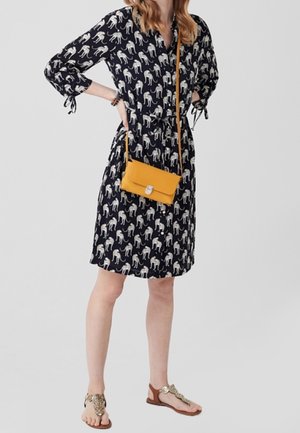 Kobieta w czarnej sukience w białe zwierzęce wzory, trzymająca musztardowożółtą torebkę na ramię i nosząca brązowe sandały z ozdobnymi paskami.