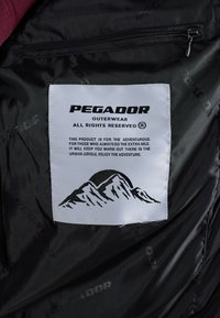 Černý textilní interiér s výraznou bílou cedulkou obsahující "PEGADOR OUTERWEAR" a grafiku hory. Hladký povrch, přítomná kapsa na zip.