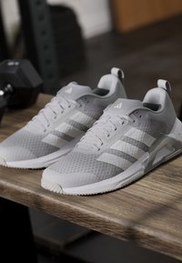 Paire de chaussures de sport Adidas gris clair avec des bandes blanches placée sur une surface en bois près d'un haltère noir.