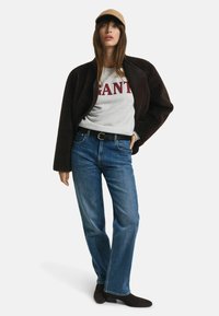 Bruine fleece jas, grijs sweatshirt met rode "GANT" tekst, blauwe rechte jeans en donkerbruine enkellaarzen; draagt een beige pet.