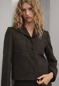 Blazer de mezcla de lana marrón con cuello redondeado, cuatro bolsillos frontales y botones negros; corte entallado y tejido texturizado.