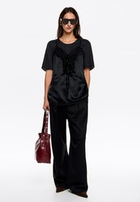 Czarny satynowy top z koronkowymi detalami nałożony na koszulę z krótkim rękawem, w połączeniu z luźnymi czarnymi spodniami i bordową skórzaną torbą.
