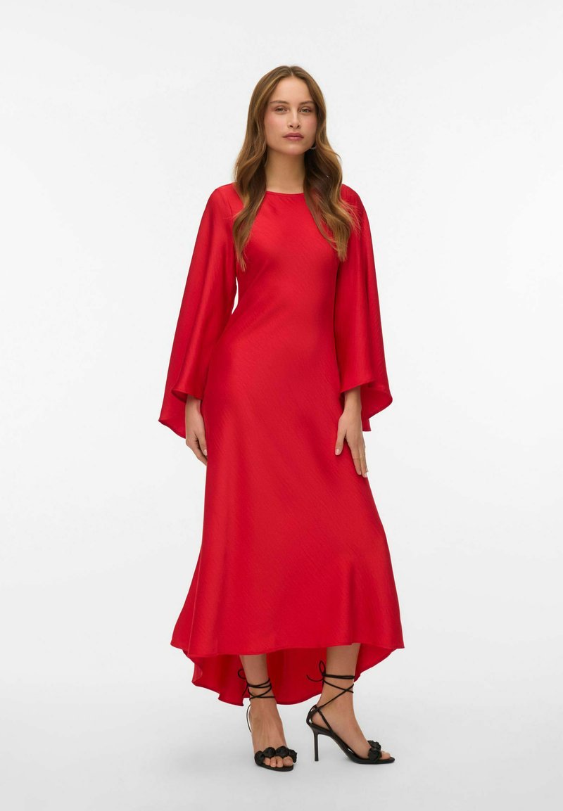 Sarkans asimetrisks kleita ar garām, plūstošām piedurknēm, gludu tekstūru un ceļgala garuma apakšdaļu priekšā, apvienota ar melnām augstpapēžu sandālēm.