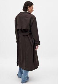 Brun trenchcoat med ryggok och bältad midja, samt vida ärmar. Matchas med ljusblå utsvängda jeans, som har en mjuk yta.