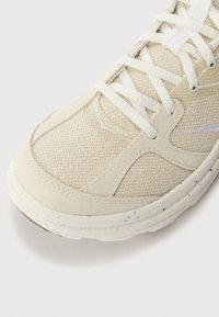 Beige athletic shoe with mesh material, textured sides, white laces, padded collar, and a speckled rubber sole.