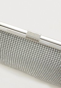 Srebrna kopertówka z teksturowaną powierzchnią, ozdobiona małymi, okrągłymi ćwiekami. Metalowy zamek z napisem "MASCARA" uzupełnia całość.