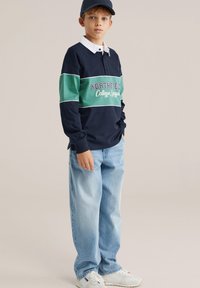 Tummansininen pitkähihaisen poolopaita, jossa vihreä raita ja teksti "NORTHFIELD College League", yhdistetty vaaleansinisiin leveisiin farkkuihin ja valkoisiin lenkkareihin.