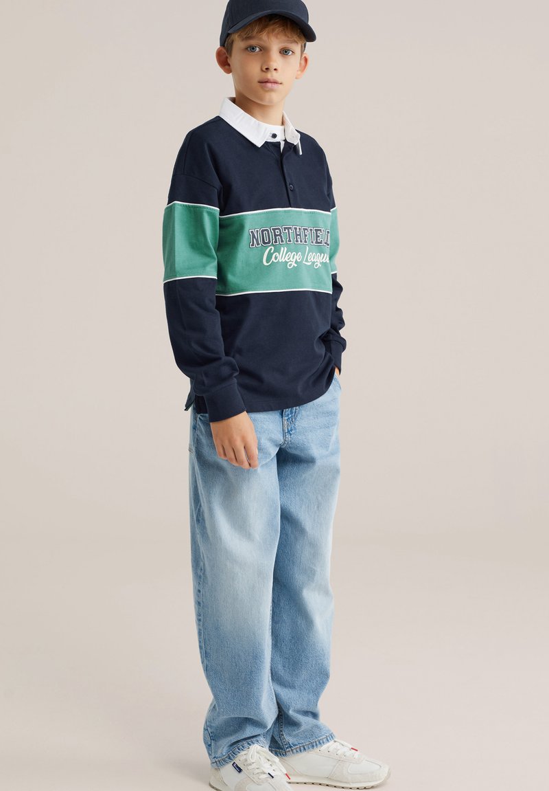 Tummansininen pitkähihaisen poolopaita, jossa vihreä raita ja teksti "NORTHFIELD College League", yhdistetty vaaleansinisiin leveisiin farkkuihin ja valkoisiin lenkkareihin.