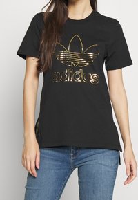 Svart bomull t-shirt med kort ärm som har en glänsande guld Adidas-logotyp och trefoil-design på framsidan. Avslappnad passform, utan dekorationer.