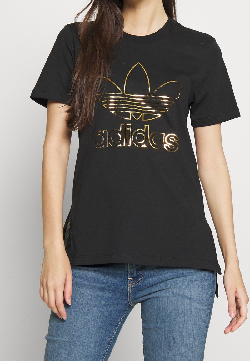 Svart bomull t-shirt med kort ärm som har en glänsande guld Adidas-logotyp och trefoil-design på framsidan. Avslappnad passform, utan dekorationer.