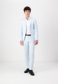 Világoskék, slim fit öltöny, egy gombos zakóval és hozzávaló nadrággal. Fehér inggel és fekete bőr cipővel párosítva.