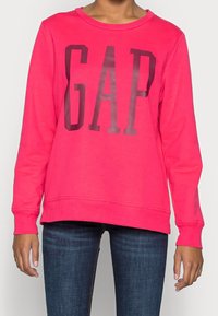 Felpa rosa con logo "GAP" in stampa scura. Presenta maniche lunghe, collo rotondo e polsini a costine. Il materiale sembra morbido ed elasticizzato.