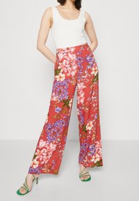 Kwiatowe spodnie z szerokimi nogawkami w kolorze czerwonym, ozdobione intensywnymi różowymi, fioletowymi i zielonymi kwiatami. W zestawie z białą, prążkowaną bluzką i zielonymi sandałami na obcasie.