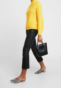 Pull jaune à torsades, pantalon court en cuir noir, sac à main noir avec ferrures dorées et chaussures slip-on à motif noir et blanc.