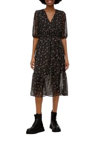 Kvinna klädd i en svart blommig midiklänning med puffärmar och svarta snörboots, leende, stående framför en vit bakgrund.