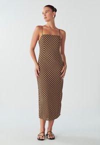 Žena stojaca v hnedo-čiernych geometrických vzorovaných midi šatách s tenkými ramienkami a čiernymi sandálmi na jednotnom bielom pozadí.