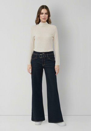 Frau mit hellbeigem Rollkragenpullover und hoch taillierten, dunklen, weiten Jeans mit Gürtel, die vor einem schlichten hellen Hintergrund steht.