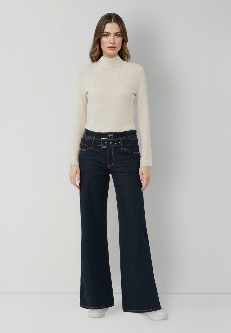 Kobieta w jasnobeżowym swetrze z golfem i ciemnych, szerokich spodniach z wysokim stanem, z paskiem, stojąca na tle jasnej, jednolitej ściany.