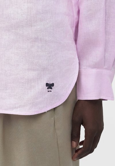 Ανοιχτό ροζ λινό πουκάμισο με μικρή μαύρη κεντημένη πεταλούδα και μπεζ παντελόνι, εστίαση στο τελείωμα και την μανσέτα του πουκαμίσου κοντά σε σκούροχρωμο χέρι.