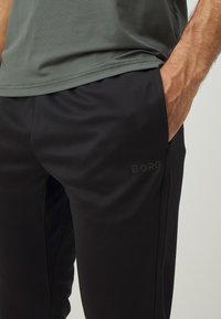 Fekete sportnadrág sima textúrával, oldalzsebekkel és egy diszkrét "BORG" logóval a bal combon. Szürke felsővel párosítva.