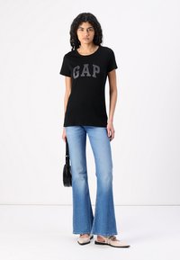 Zwarte katoenen t-shirt met het "GAP" logo in grijs, gecombineerd met lichtblauwe uitlopende jeans. Het model houdt een kleine zwarte handtas vast.