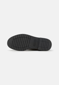 Black rubber shoe sole with horizontal and vertical tread patterns, featuring a logo stamp and slight curvature for ergonomic support.