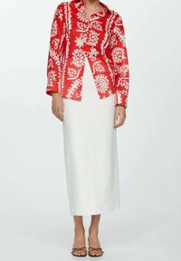 Persoon in een rood en wit gestreepte blouse met knopen, een witte midi-jurk en bruine sandaaltjes met bandjes, staande tegen een effen achtergrond.