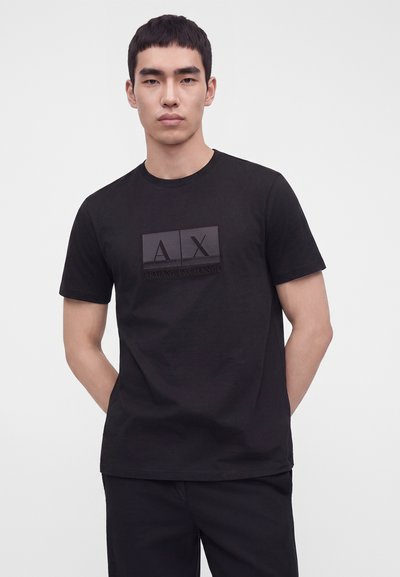 Млад мъж с черна тениска Armani Exchange с фин изображен логотип, застанал с ръце зад гърба пред обикновен фон.