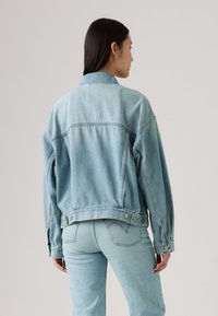 Veste en denim bleu clair avec une coupe décontractée, des manches surdimensionnées et des accents de boutons aux poignets et à la taille. Dispose d'une couture en y à l'arrière.