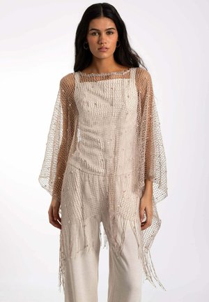 Châle en tricot beige avec un tissage ouvert, doté de larges manches et de bords frangés, superposé sur une tenue texturée de couleur claire.