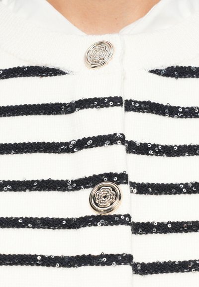 Vit cardigan med svarta horisontella ränder och paljettdetaljer, rund silverblomknapp och en texturerad stickad tyg.