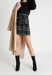 Blusa nera a maniche lunghe con dettagli in pizzo, mini gonna a quadri con tasche, trench beige e stivaletti neri alla caviglia.