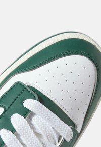 Scarpa da ginnastica in pelle verde e bianca con finitura testurizzata, caratterizzata da perforazioni, colletto imbottito e lacci bianchi. Punta arrotondata, suola flessibile.