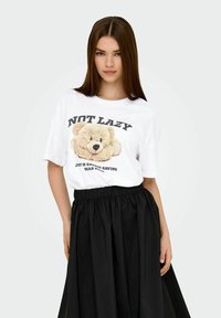 White T-shirt with a teddy bear graphic and black text; paired with a black, flowy skirt with an elastic waistband. Soft material and relaxed fit.
