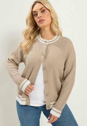 Vrouw met blond golvend haar die een beige vest met knopen draagt met witte gestreepte manchetten en kraag over een wit overhemd en blauwe spijkerbroek.