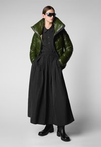 Gewatteerde groene jas met hoge kraag, zwart button-up shirt en lange zwarte rok. Zwarte enkellaarzen maken de outfit af.