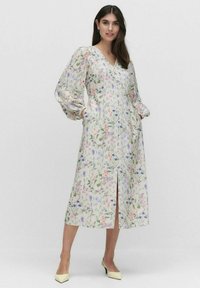 Kukallinen mekko V-pääkohdalla, puffeilla hihoilla ja sivutaskuilla. Kevyt kangas esittelee vaaleita kukkakuosisia kuvioita kermanvärisellä taustalla. Keskikorkeus sääriin.