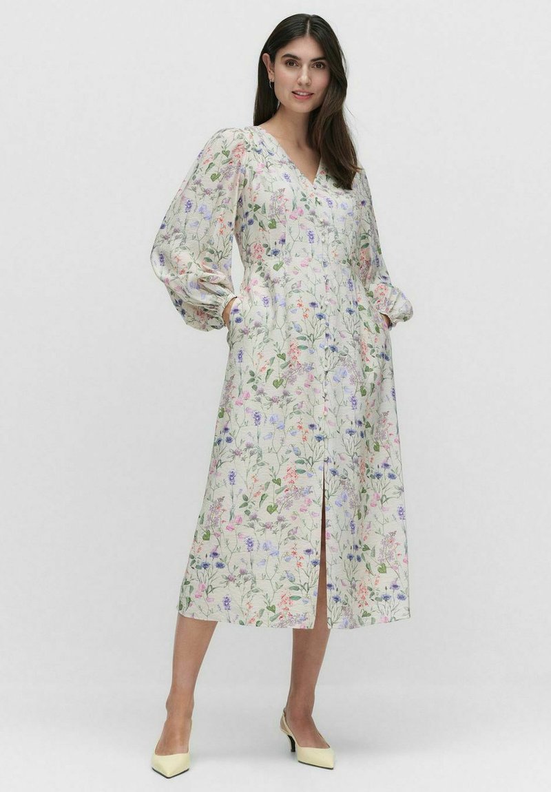 Kukallinen mekko V-pääkohdalla, puffeilla hihoilla ja sivutaskuilla. Kevyt kangas esittelee vaaleita kukkakuosisia kuvioita kermanvärisellä taustalla. Keskikorkeus sääriin.