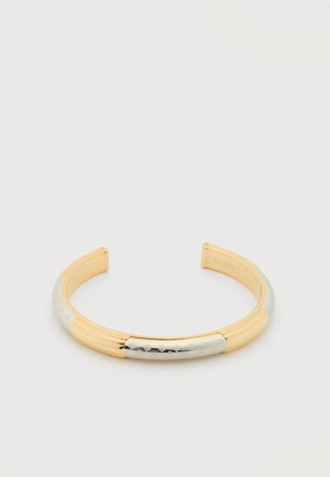 Open armband van goud en zilver met gegraveerde patronen en geribbelde secties, aan de binnenkant gebrandmerkt met "Guess".