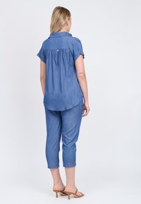 Kurzärmeliges blaues Chambray-Hemd mit abgerundetem Saum, ausgestattet mit Rückenfalten und einem kleinen goldenen Knopf. Kombiniert mit passenden blauen, verkürzten Hosen.