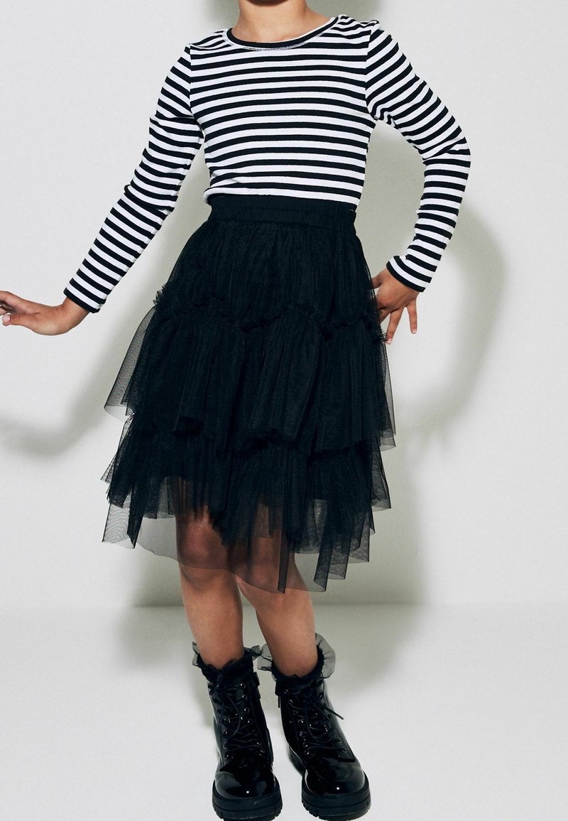Enfant portant une jupe en tulle noire à volants, des bottes noires brillantes à lacets et un t-shirt à manches longues rayé noir et blanc, debout les bras légèrement levés.