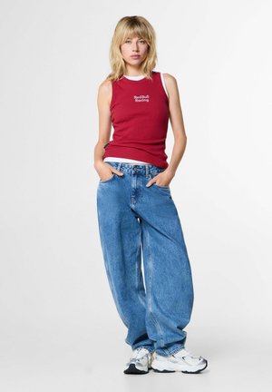 Jonge vrouw met blond haar die een mouwloos rood Red Bull Racing top, wijde blauwe spijkerbroek en witte sneakers draagt, staand met handen in de zakken.